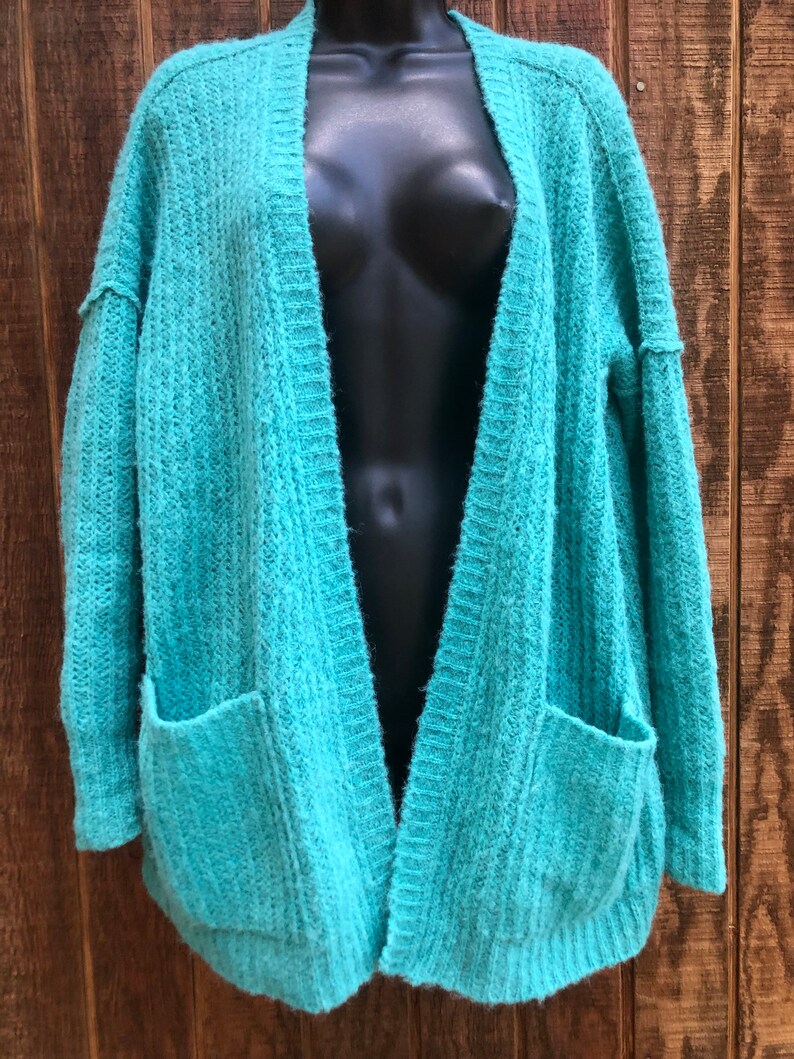 May include: A teal blue open-front cardigan with long sleeves and two patch pockets. The cardigan is made of a soft, fuzzy knit fabric.