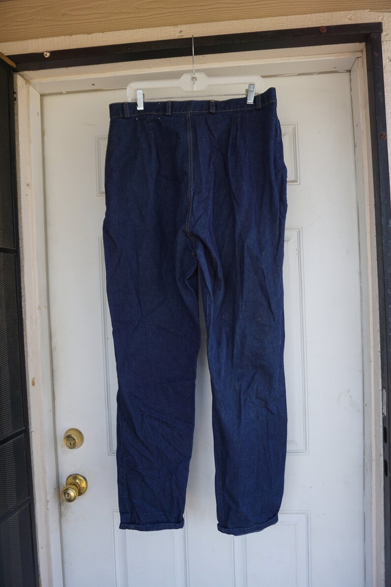 May include: Dark blue denim trousers hanging on a white hanger. The trousers are a solid colour with a classic five-pocket design. The trousers are hanging on a white door.
