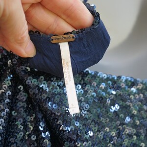 K&ouml;nnte beinhalten: Nahaufnahme eines marineblauen Kleidungsst&uuml;cks mit Pailletten. Das Kleidungsst&uuml;ck hat ein dunkelblaues Futter und ein Etikett mit der Aufschrift "Free People" und "MADE IN INDIA". Die Pailletten sind eine Mischung aus Blau und Silber.