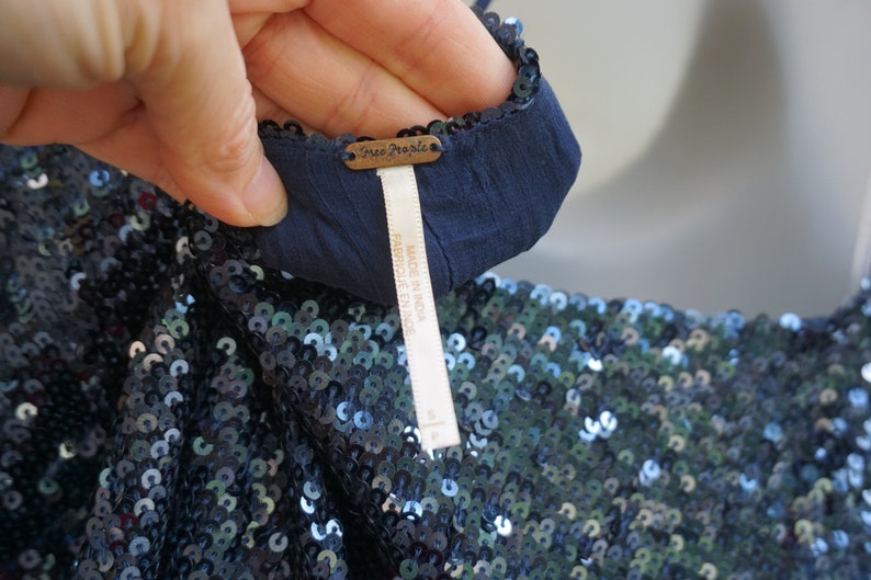 K&ouml;nnte beinhalten: Nahaufnahme eines marineblauen Kleidungsst&uuml;cks mit Pailletten. Das Kleidungsst&uuml;ck hat ein dunkelblaues Futter und ein Etikett mit der Aufschrift "Free People" und "MADE IN INDIA". Die Pailletten sind eine Mischung aus Blau und Silber.