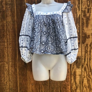 Puede incluir: Una blusa estampada en azul y blanco con mangas largas y cuello cuadrado. La blusa presenta un dise&ntilde;o geom&eacute;trico en el corpi&ntilde;o y estampados florales en las mangas. Las mangas tienen un detalle de volantes en el hombro y est&aacute;n ribeteadas con cinta negra.