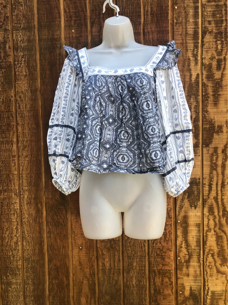 Puede incluir: Una blusa estampada en azul y blanco con mangas largas y cuello cuadrado. La blusa presenta un dise&ntilde;o geom&eacute;trico en el corpi&ntilde;o y estampados florales en las mangas. Las mangas tienen un detalle de volantes en el hombro y est&aacute;n ribeteadas con cinta negra.