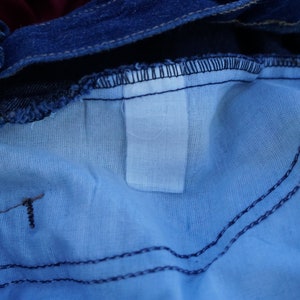May include: Close-up of the inside of blue denim jeans. The image shows the light blue lining, the dark blue denim, and the stitching details. A white label is visible.