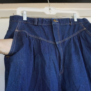May include: Dark blue denim trousers with a high waist and pleated front. The trousers have a button closure, pockets, and light-coloured stitching. The trousers are hanging on a white plastic hanger.