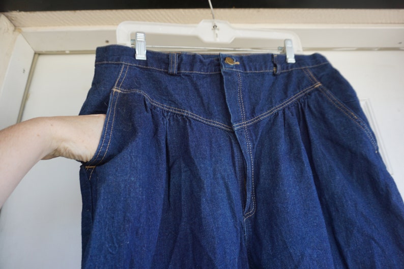May include: Dark blue denim trousers with a high waist and pleated front. The trousers have a button closure, pockets, and light-coloured stitching. The trousers are hanging on a white plastic hanger.