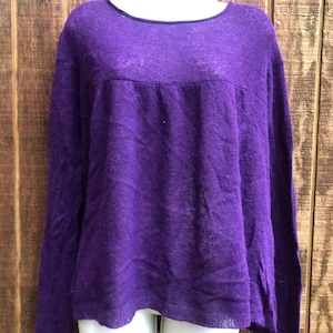 May include: A long-sleeved, purple sweater with a scoop neck. The sweater is displayed on a white mannequin. The fabric has a slightly textured appearance. The sweater's design includes a gathered detail across the upper chest area.