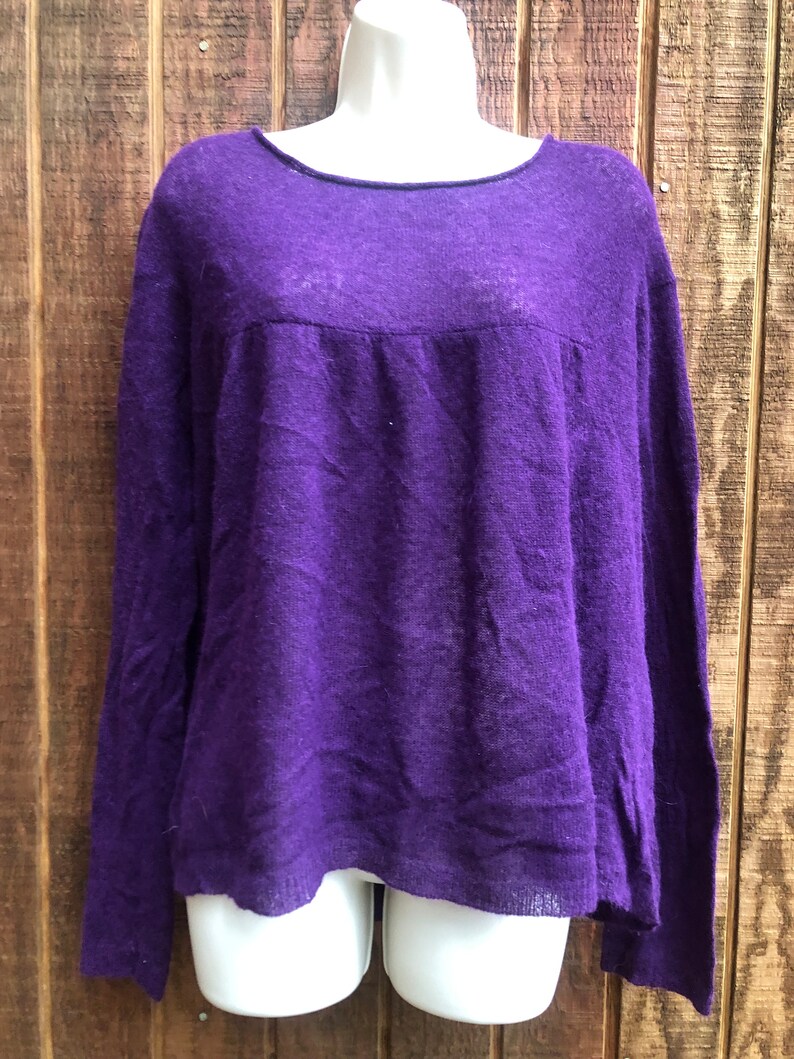 May include: A long-sleeved, purple sweater with a scoop neck. The sweater is displayed on a white mannequin. The fabric has a slightly textured appearance. The sweater's design includes a gathered detail across the upper chest area.