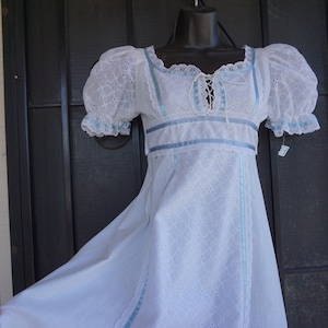 May include: White eyelet dress with blue ribbon accents. The dress features short, puffed sleeves, a lace-up bodice, and a full skirt. The dress is displayed on a black mannequin against a neutral background. The dress has a vintage, romantic style.