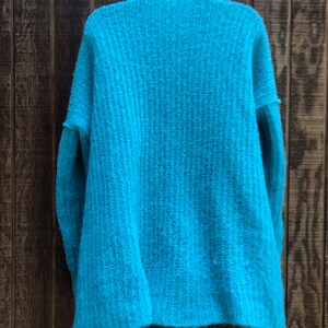 May include: A teal blue knitted cardigan with a ribbed texture. The cardigan is hanging on a wooden hanger against a wood panel background.