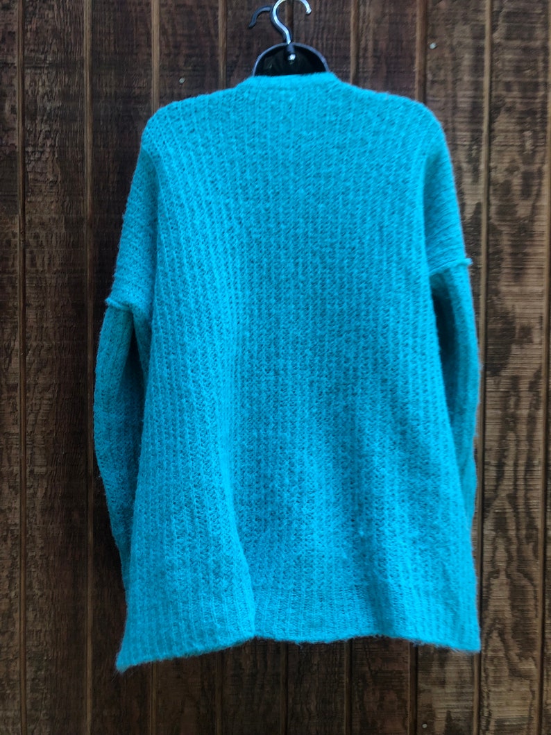 May include: A teal blue knitted cardigan with a ribbed texture. The cardigan is hanging on a wooden hanger against a wood panel background.