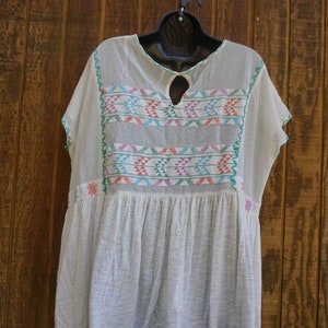 May include: A white, loose-fitting dress with colorful embroidered details. The dress features a keyhole neckline and short sleeves. The embroidery includes geometric patterns in shades of pink, blue, and green. The dress is hanging on a black hanger.