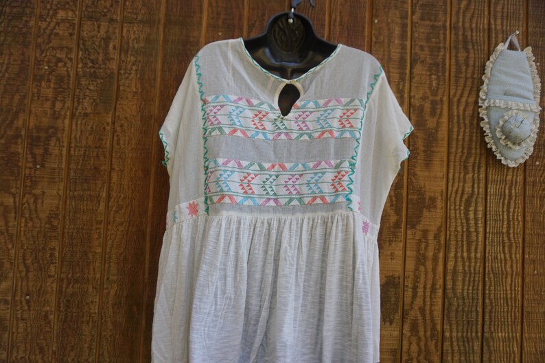 May include: A white, loose-fitting dress with colorful embroidered details. The dress features a keyhole neckline and short sleeves. The embroidery includes geometric patterns in shades of pink, blue, and green. The dress is hanging on a black hanger.