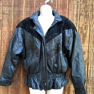 May include: Black leather jacket with a zip-up front, featuring a black suede yoke and decorative embroidery. The jacket has a cinched waist and a ruffled hem. The jacket is displayed on a white mannequin against a wooden background.
