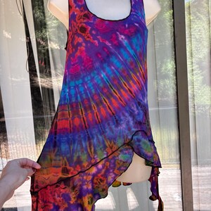 May include: A sleeveless tank top featuring a vibrant tie-dye pattern in shades of purple, blue, red, and orange. The garment has a scoop neckline and an asymmetrical, layered hemline with black stitched edges throughout the entire piece.