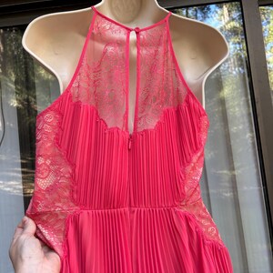 Puede incluir: Un vestido rosa brillante con una falda plisada y un cuello halter. El corpi&ntilde;o presenta delicados detalles de encaje en los hombros y la espalda. El vestido est&aacute; colgado en un maniqu&iacute; blanco.