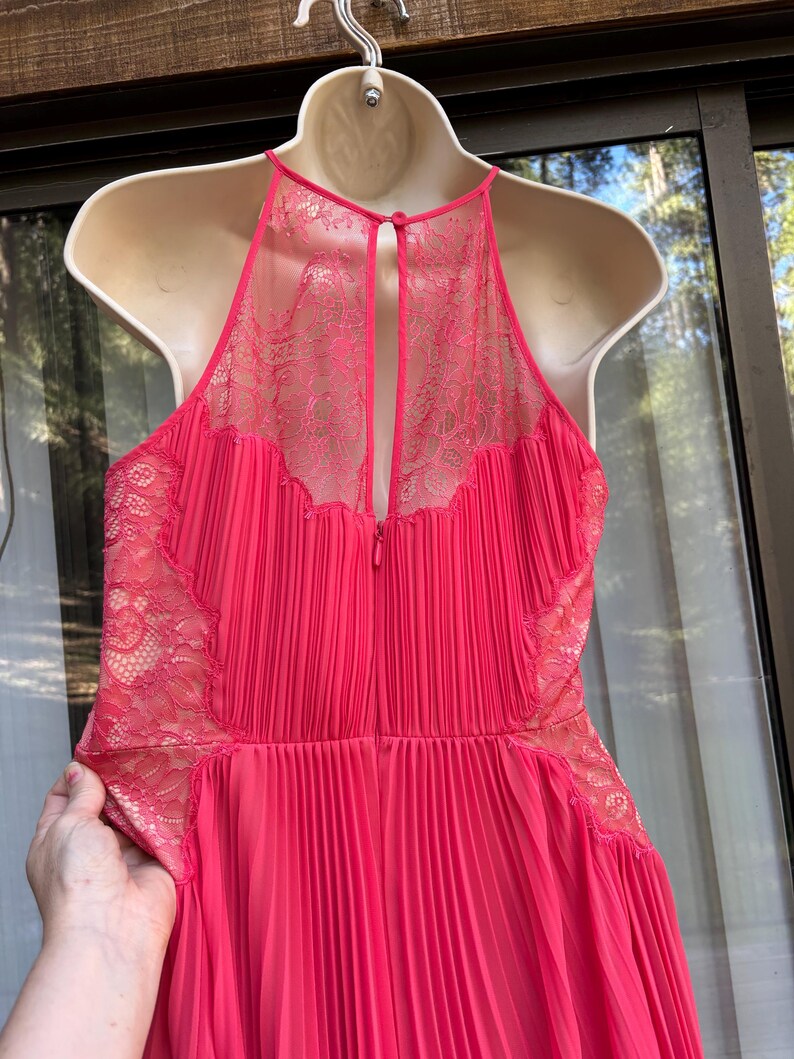 Puede incluir: Un vestido rosa brillante con una falda plisada y un cuello halter. El corpi&ntilde;o presenta delicados detalles de encaje en los hombros y la espalda. El vestido est&aacute; colgado en un maniqu&iacute; blanco.