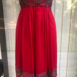 K&ouml;nnte beinhalten: Ein leuchtend rotes Kleid mit einem bestickten Oberteil und Saum. Das Kleid hat einen flie&szlig;enden, knielangen Rock und einen detaillierten, verzierten G&uuml;rtel. Die Perlen enthalten silberne und dunkle Akzente, die der Abendmode einen Hauch von Eleganz verleihen.