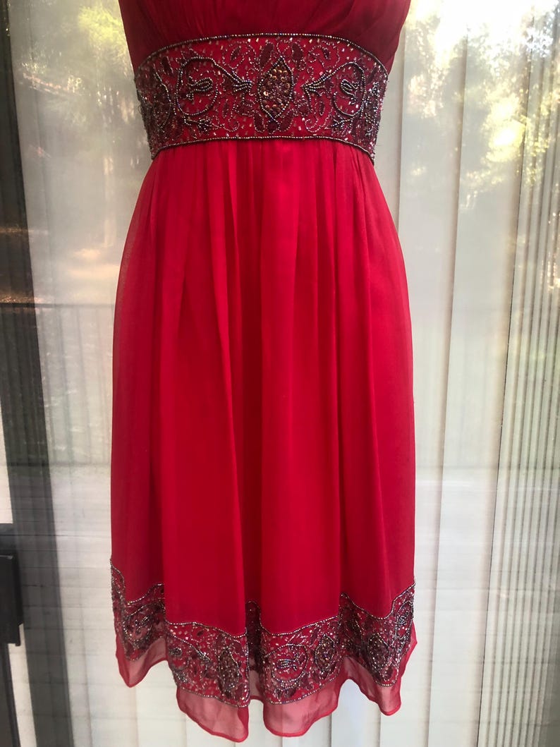 K&ouml;nnte beinhalten: Ein leuchtend rotes Kleid mit einem bestickten Oberteil und Saum. Das Kleid hat einen flie&szlig;enden, knielangen Rock und einen detaillierten, verzierten G&uuml;rtel. Die Perlen enthalten silberne und dunkle Akzente, die der Abendmode einen Hauch von Eleganz verleihen.