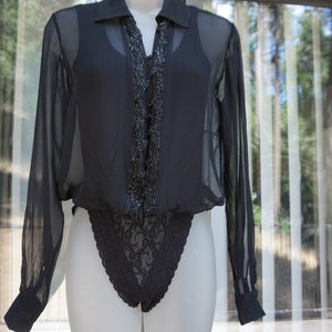 May include: Black sheer bodysuit with long sleeves and a collared neckline. The front features a beaded embellishment and a lace bottom. The garment is displayed on a white mannequin against a backdrop of a window with vertical blinds.