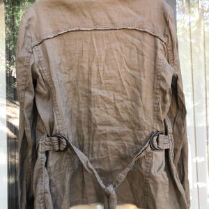 K&ouml;nnte beinhalten: Eine beige Leinenjacke mit ausgefranstem Kragen und Saum. Die Jacke hat einen G&uuml;rtel mit zwei schwarzen Metallschnallen und einem Bindverschluss in der Taille. Die R&uuml;ckseite der Jacke ist sichtbar und zeigt das Design und die N&auml;hte.