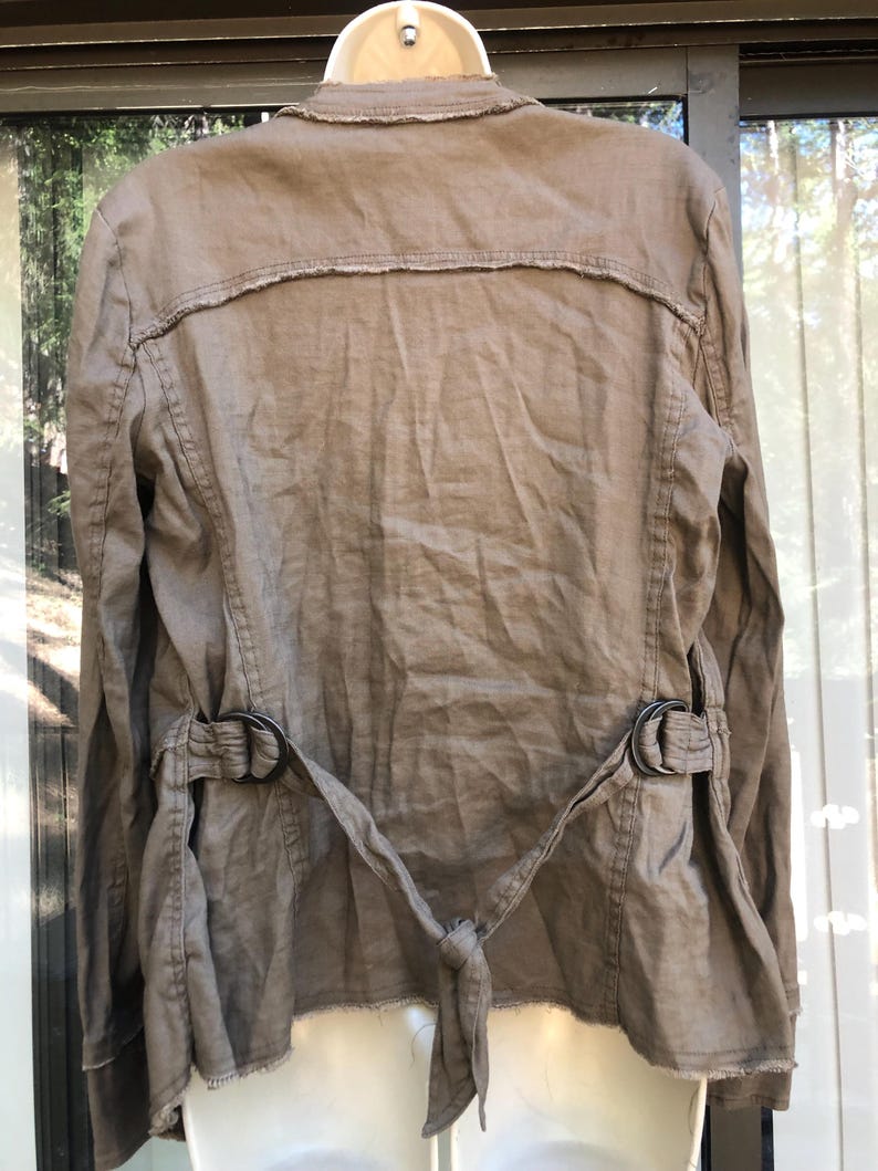 K&ouml;nnte beinhalten: Eine beige Leinenjacke mit ausgefranstem Kragen und Saum. Die Jacke hat einen G&uuml;rtel mit zwei schwarzen Metallschnallen und einem Bindverschluss in der Taille. Die R&uuml;ckseite der Jacke ist sichtbar und zeigt das Design und die N&auml;hte.