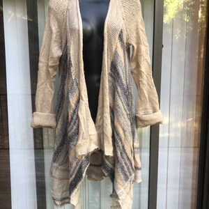 May include: A beige knitted cardigan with a long, asymmetrical hemline. The cardigan features a blue and beige striped pattern and a loose, open weave.