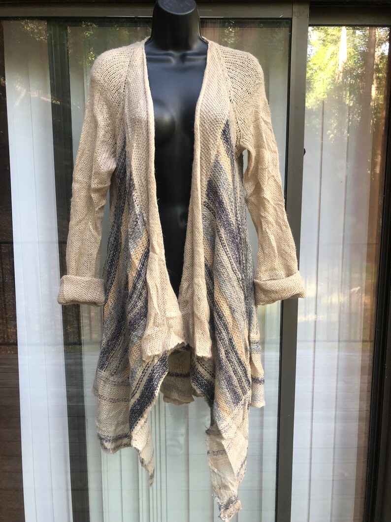 May include: A beige knitted cardigan with a long, asymmetrical hemline. The cardigan features a blue and beige striped pattern and a loose, open weave.