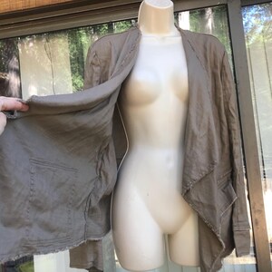 K&ouml;nnte beinhalten: Offene Jacke in Taupe mit zwei Vordertaschen und einem rohen Saum. Die Jacke wird auf einer Schaufensterpuppe pr&auml;sentiert. Der Stoff scheint eine leichte Leinen- oder Baumwollmischung zu sein. Die Jacke hat eine neutrale Farbe.