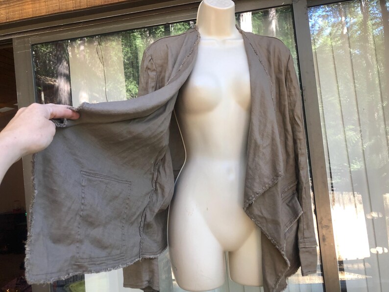 K&ouml;nnte beinhalten: Offene Jacke in Taupe mit zwei Vordertaschen und einem rohen Saum. Die Jacke wird auf einer Schaufensterpuppe pr&auml;sentiert. Der Stoff scheint eine leichte Leinen- oder Baumwollmischung zu sein. Die Jacke hat eine neutrale Farbe.