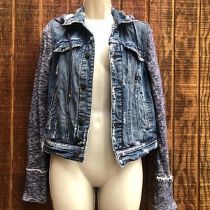 May include: A blue denim jacket with a distressed look, featuring a hood and sleeves made of a heathered gray knit fabric. The jacket has button closures and pockets, displayed on a mannequin.