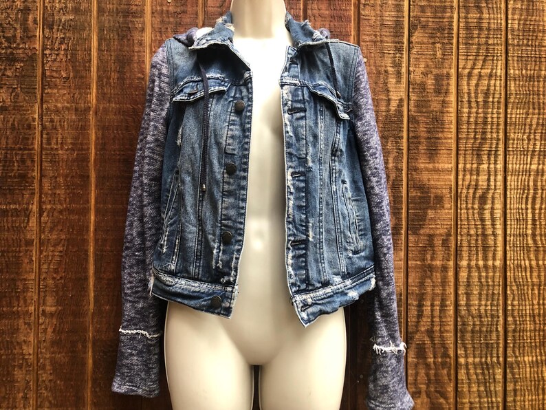 May include: A blue denim jacket with a distressed look, featuring a hood and sleeves made of a heathered gray knit fabric. The jacket has button closures and pockets, displayed on a mannequin.