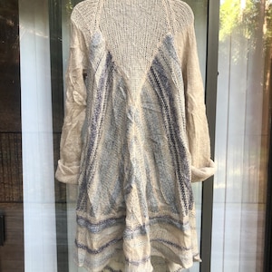 May include: A beige knitted cardigan with a blue and white striped pattern. The cardigan has a long, asymmetrical hemline and a loose fit.