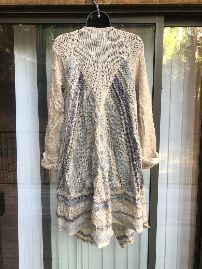 May include: A beige knitted cardigan with a blue and white striped pattern. The cardigan has a long, asymmetrical hemline and a loose fit.