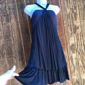 Op de afbeelding: Een marineblauwe halterjurk met een ringdetail bij de halslijn. De jurk heeft een zwierige rok met een gefranjerde zoom. De jurk wordt getoond op een witte mannequin tegen een houten achtergrond.
