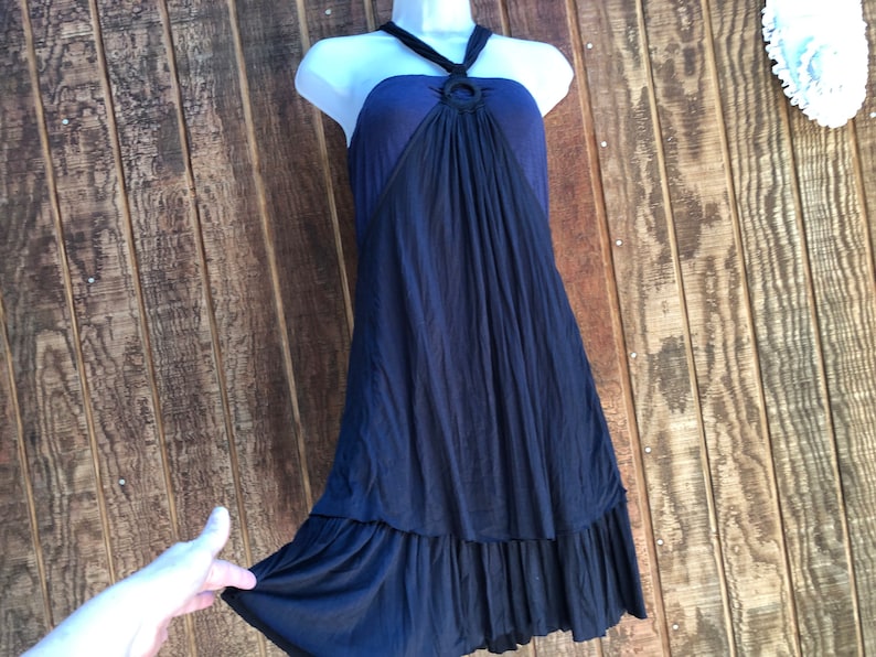 Op de afbeelding: Een marineblauwe halterjurk met een ringdetail bij de halslijn. De jurk heeft een zwierige rok met een gefranjerde zoom. De jurk wordt getoond op een witte mannequin tegen een houten achtergrond.