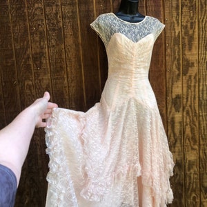 Puede incluir: Vestido vintage color melocotón con escote corazón y delicada superposición de encaje. El vestido presenta un corpiño ajustado y una falda fluida con ribete de encaje con volantes. Exhibido en un maniquí negro sobre fondo de madera.