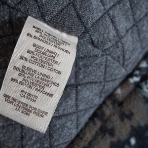 May include: Close-up of a white clothing tag with black text, detailing fabric composition: polyester, spandex, cotton, and rayon. The tag is against a gray quilted fabric background.