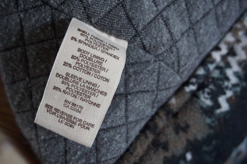 May include: Close-up of a white clothing tag with black text, detailing fabric composition: polyester, spandex, cotton, and rayon. The tag is against a gray quilted fabric background.