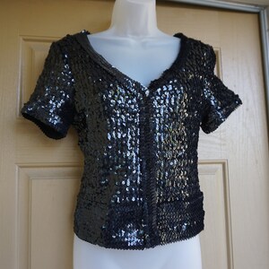 May include: A black sequined top with short sleeves and a V-neck. The top is covered in small, shiny sequins, giving it a glamorous appearance. The garment is displayed on a white mannequin, showcasing its design and fit.