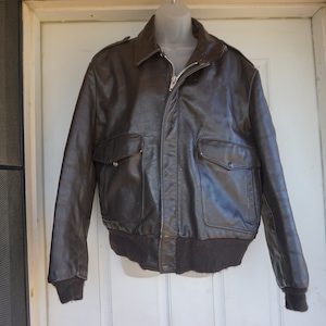 May include: A brown leather bomber jacket with a zip-up front, two front pockets, and a ribbed hem and cuffs. The jacket is displayed on a mannequin. The jacket's style is reminiscent of vintage aviation apparel.