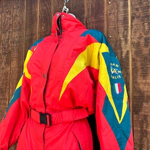 May include: A vintage red ski suit with yellow and teal detailing. The suit has a black belt, a high collar, and the text "TEAM ITALIA" with an Italian flag patch. It hangs against a wooden backdrop.