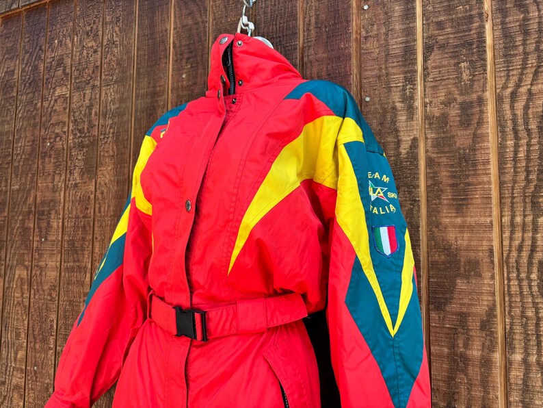 May include: A vintage red ski suit with yellow and teal detailing. The suit has a black belt, a high collar, and the text "TEAM ITALIA" with an Italian flag patch. It hangs against a wooden backdrop.