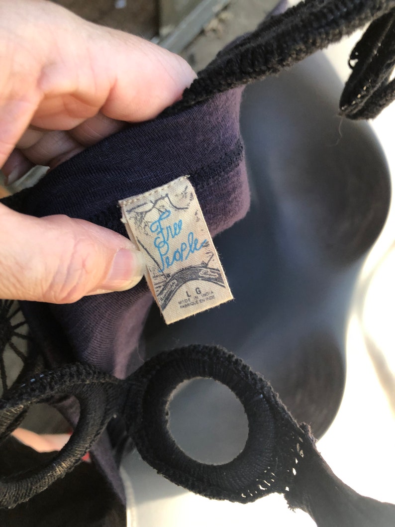 Op de afbeelding: Close-up van een donkerpaarse top met zwarte bandjes. Het label leest "Free People" in lichtblauw schrift. Het label geeft ook de maat Large en Made in India aan.