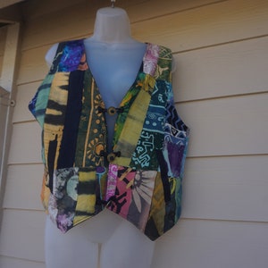 May include: A colorful patchwork vest with a V-neck design. The vest features a variety of patterns and colors, including blue, green, yellow, and pink. It has two wooden buttons for closure and is displayed on a mannequin.