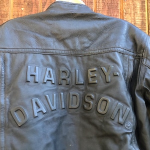 May include: Black leather jacket with the embossed text "HARLEY-DAVIDSON" across the back. The jacket has a zipper on the left side and a classic biker style. The leather appears worn, giving it a vintage look.