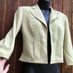 May include: A light green blazer with a notched collar and long sleeves. The jacket has a tailored fit with vertical seams and a split cuff detail. The blazer is displayed on a black mannequin against a wooden background.