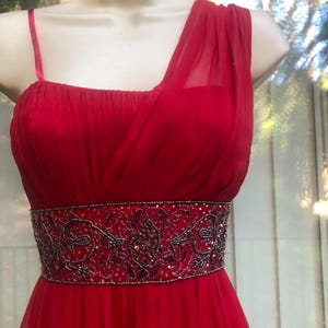 K&ouml;nnte beinhalten: Ein rotes Abendkleid mit One-Shoulder-Design und einem bestickten G&uuml;rtel. Das Kleid hat einen flie&szlig;enden, bodenlangen Rock und ein eng anliegendes Oberteil. Der G&uuml;rtel ist mit aufwendigen Perlenarbeiten in Rot-, Schwarz- und Goldt&ouml;nen verziert.