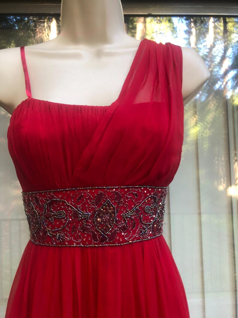 K&ouml;nnte beinhalten: Ein rotes Abendkleid mit One-Shoulder-Design und einem bestickten G&uuml;rtel. Das Kleid hat einen flie&szlig;enden, bodenlangen Rock und ein eng anliegendes Oberteil. Der G&uuml;rtel ist mit aufwendigen Perlenarbeiten in Rot-, Schwarz- und Goldt&ouml;nen verziert.