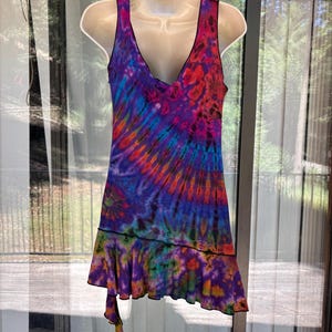 May include: A sleeveless dress featuring a vibrant tie-dye pattern in shades of purple, blue, orange, and red. The garment has a deep V-neckline, a fitted bodice, and a ruffled hemline that creates an asymmetrical, layered effect at the bottom edge.