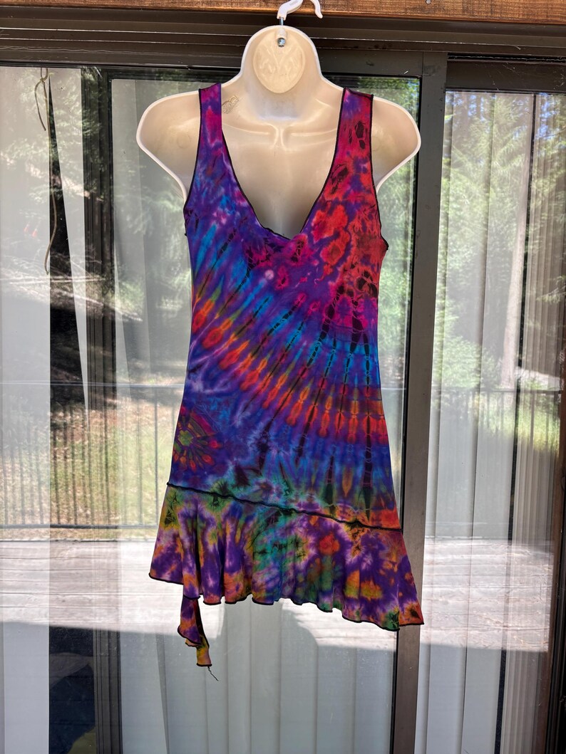 May include: A sleeveless dress featuring a vibrant tie-dye pattern in shades of purple, blue, orange, and red. The garment has a deep V-neckline, a fitted bodice, and a ruffled hemline that creates an asymmetrical, layered effect at the bottom edge.